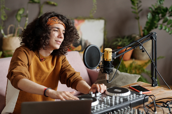 A guy with dark curls in a bandana dressed in a sweatshirt is sitting at a table with DJ equipment on it and using it sings into a microphone with a pop filter